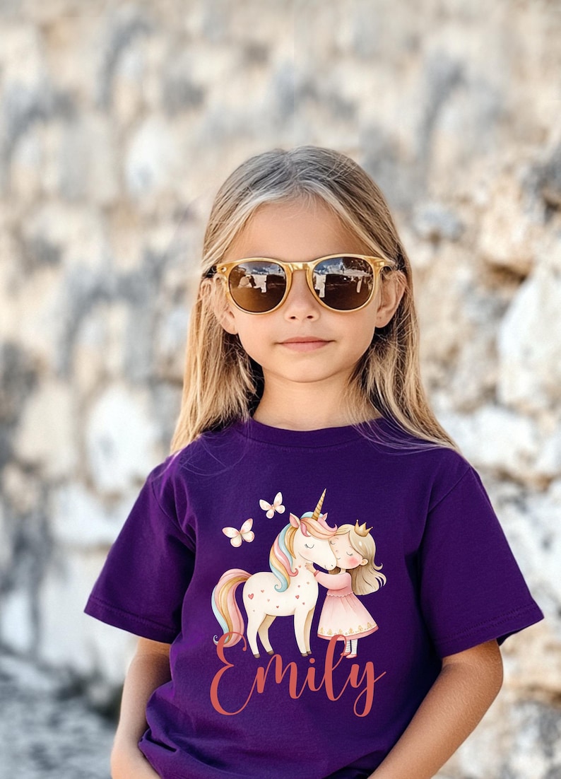 Pu&ograve; includere: Una maglietta viola per bambini con un disegno di unicorno e principessa, con il nome "Emily" in corsivo. La persona indossa occhiali da sole con montatura dorata. Sfondo strutturato.