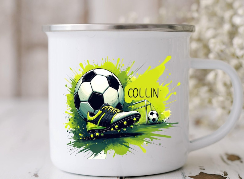 May include: White enamel mug featuring a soccer theme. The design includes a soccer ball, cleats, and a small goal, set against a backdrop of green and yellow paint splatters. The name "COLLIN" is also displayed.