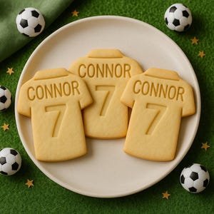 以下が含まれることがあります： 白いお皿の上に、名前「CONNOR」と数字「7」が入った、黄色のサッカーユニフォーム型のクッキーが3つ並んでいます。ミニチュアサッカーボールと金色の星型のスプリンクルが、緑色の表面にあるお皿を囲んでいます。