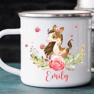 以下が含まれることがあります： 白地にエナメルコーティングされたマグカップ。茶色と白の馬のイラスト、ピンクの花、緑の葉が描かれている。マグカップは銀色の縁と持ち手がある。マグカップの下部に赤い文字で「Emily」と書いてある。