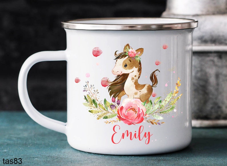 以下が含まれることがあります： 白地にエナメルコーティングされたマグカップ。茶色と白の馬のイラスト、ピンクの花、緑の葉が描かれている。マグカップは銀色の縁と持ち手がある。マグカップの下部に赤い文字で「Emily」と書いてある。