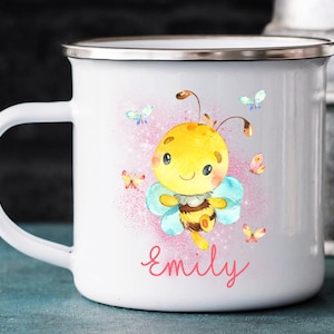 Puede incluir: Taza de esmalte blanco con un fondo rosa y blanco que presenta una abeja de dibujos animados con alas azules y el nombre "Emily" en letras rosas.