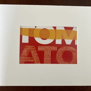 Op de afbeelding: Een ingelijste kunstdruk met het woord "TOMATO" in grote witte letters op een rode achtergrond. De letters zijn bedekt met een textuurpatroon. Een horizontale oranje streep loopt over de bovenkant. De print is in een witte passe-partout.
