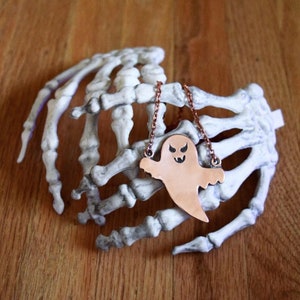 Könnte beinhalten: Eine kupferfarbene Geister-Anhänger-Kette mit einer Kette, die an einer weißen Plastik-Skeletthand hängt.