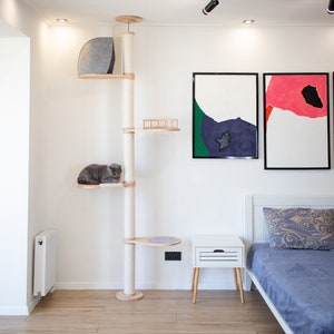 May include: A white cat tree with a gray cat bed, a gray cat sitting on a platform, and a round platform with a gray surface. The cat tree is in a room with white walls, a white bed with a blue blanket, a white nightstand with a drawer, and two framed abstract paintings on the wall.