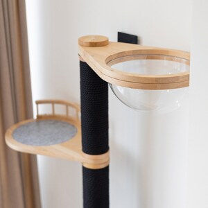 May include: A modern cat tree with a light wood frame and black rope-wrapped post. Features a clear, spherical cat bed and a small, round platform with a gray felt pad. The cat tree is mounted on a white wall.