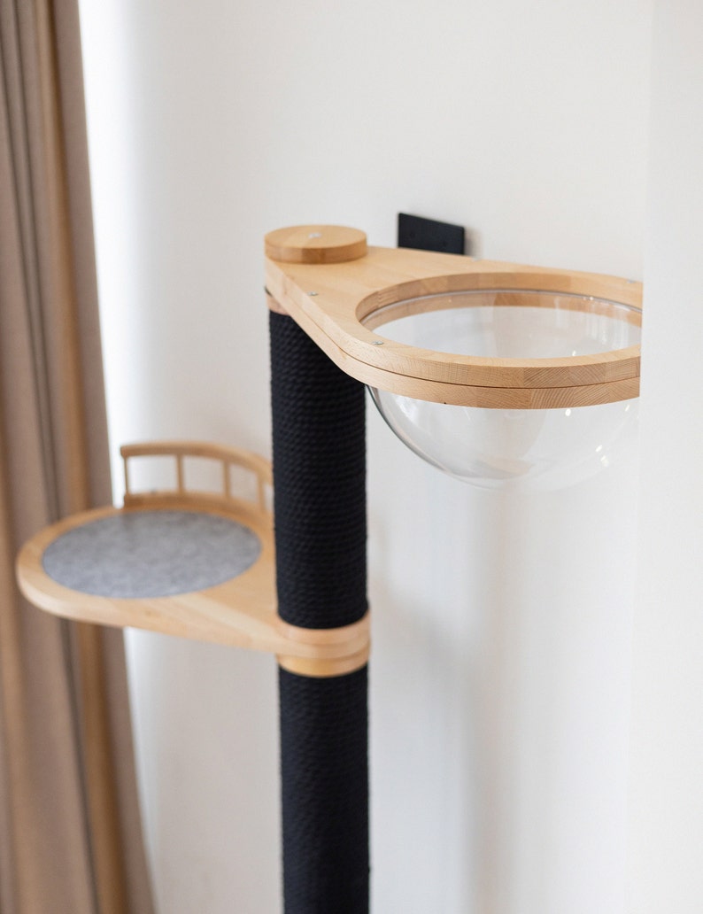 May include: A modern cat tree with a light wood frame and black rope-wrapped post. Features a clear, spherical cat bed and a small, round platform with a gray felt pad. The cat tree is mounted on a white wall.