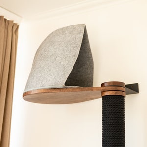 May include: A wall-mounted cat perch featuring a dark wood platform and a gray felt hood. The structure includes a vertical post wrapped in thick black rope, providing a textured surface. The unit is secured to the wall with a black metal bracket.