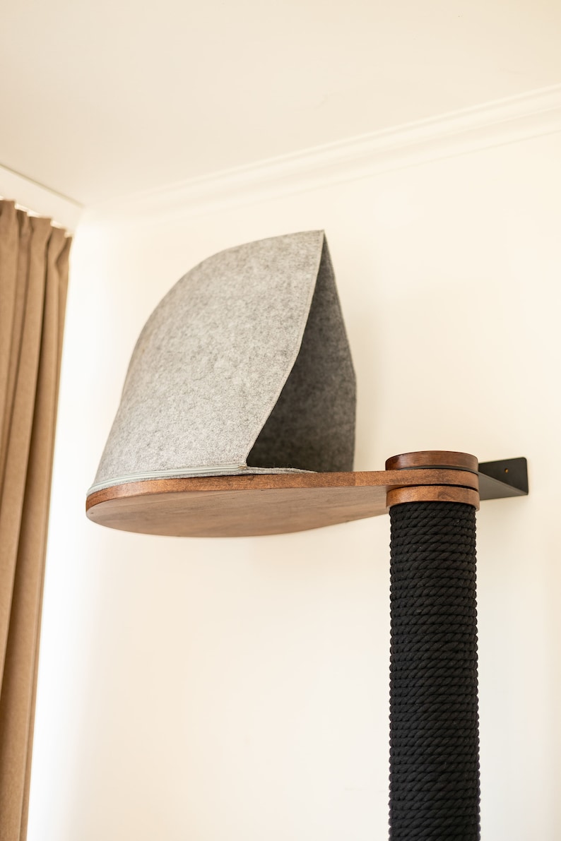 May include: A wall-mounted cat perch featuring a dark wood platform and a gray felt hood. The structure includes a vertical post wrapped in thick black rope, providing a textured surface. The unit is secured to the wall with a black metal bracket.