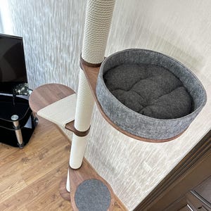 Op de afbeelding: Een kattenboom met een sisal-omwikkelde paal, houten plateaus en een grijs vilten bed. De kattenboom heeft een neutraal kleurenschema met een cr&egrave;mekleurige paal en bruine houten accenten. Het ontwerp omvat een krabpaal en een gezellig bed.