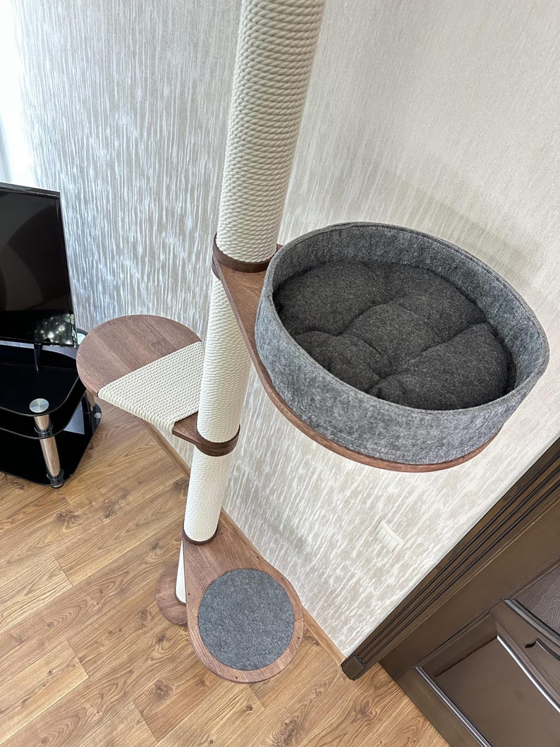 Op de afbeelding: Een kattenboom met een sisal-omwikkelde paal, houten plateaus en een grijs vilten bed. De kattenboom heeft een neutraal kleurenschema met een cr&egrave;mekleurige paal en bruine houten accenten. Het ontwerp omvat een krabpaal en een gezellig bed.
