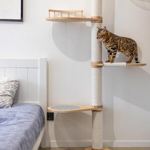 May include: A multi-level cat tree with a natural wood finish and sisal rope-wrapped posts. The cat tree features multiple platforms, including a round one with a gray felt pad. A Bengal cat stands on one of the platforms.