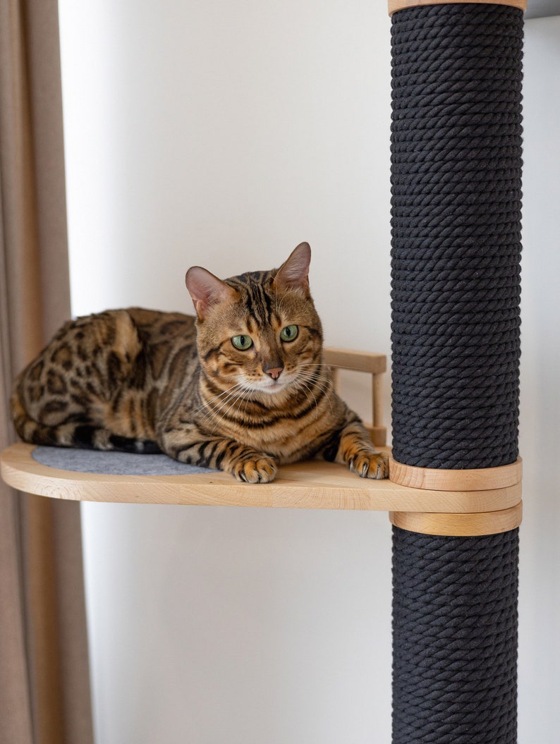 May include: A Bengal cat with brown and black spotted fur rests on a light-colored wooden platform attached to a vertical scratching post wrapped in thick, dark charcoal-colored rope. The platform features a circular gray felt inlay.