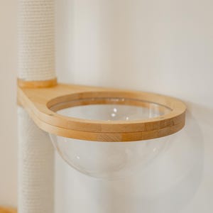 Peut inclure: Un perchoir pour chat fix&eacute; au mur avec un cadre en bois clair et un bol h&eacute;misph&eacute;rique en acrylique transparent. Le perchoir est attach&eacute; &agrave; un poteau vertical recouvert de corde de sisal blanche, offrant une structure stable pour le repos.