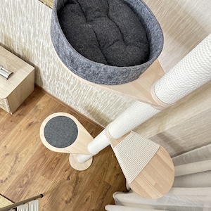 Op de afbeelding: Een kattenboom met meerdere niveaus met een grijs vilten bed, een sisal krabpaal en houten platforms. De kattenmeubels hebben een modern design en zijn gemaakt van lichtgekleurd hout en wit touw.