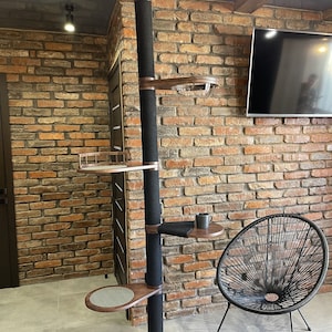 May include: A tall cat tree with a black sisal-covered post and multiple wooden platforms. The tree is against a brick wall with a black chair in the foreground.