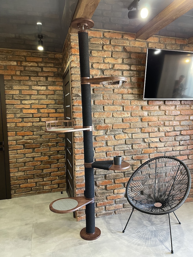 May include: A tall cat tree with a black sisal-covered post and multiple wooden platforms. The tree is against a brick wall with a black chair in the foreground.