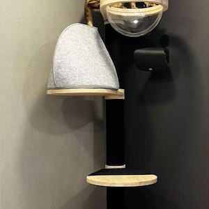 Peut inclure: Un arbre &agrave; chat &agrave; plusieurs niveaux avec un poteau central noir, des plateformes en bois clair, une cachette recouverte de feutre gris et un lit d&ocirc;me en acrylique transparent. Un chat tabby brun est assis sur la plateforme sup&eacute;rieure.