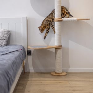 May include: A tall cat tree featuring light wood platforms and a central vertical post wrapped in thick white rope. A spotted brown Bengal cat is captured in motion, leaping from the upper level to a lower platform covered in gray fabric.
