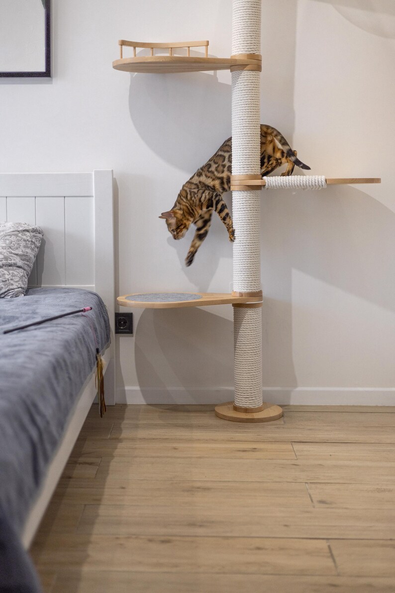 May include: A tall cat tree featuring light wood platforms and a central vertical post wrapped in thick white rope. A spotted brown Bengal cat is captured in motion, leaping from the upper level to a lower platform covered in gray fabric.