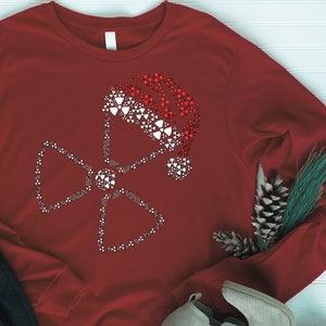 May include: A long-sleeved, claret-coloured shirt with a white and red design of a Santa hat and a radiation symbol, both made of small heart shapes. The shirt is displayed with black trousers and a pair of shoes.