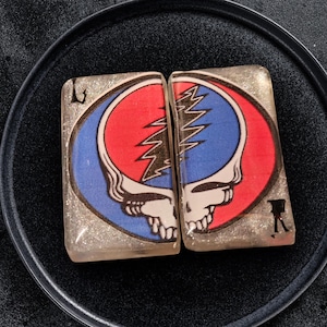 May include: Two rectangular, clear resin coasters with a Steal Your Face skull design. The design features a red and blue circle with a lightning bolt and skull. The coasters have a glittery, shimmering effect and are set on a black plate.