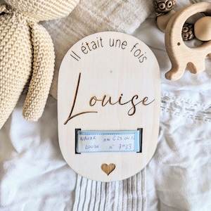 Puede incluir: Un letrero ovalado de madera con el texto "Il était une fois" y el nombre "Louise" grabado en él. Una cinta azul con el texto "NAUVAR mai l'29 ou 12 LOUISE a 7h33" está unida al letrero.