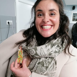 Pu&ograve; includere: Una donna indossa un cappotto bianco e una sciarpa verde con un motivo floreale e tiene in mano una piccola statuetta di legno di una donna con un vestito colorato. La statuetta &egrave; alta circa 10 cm.