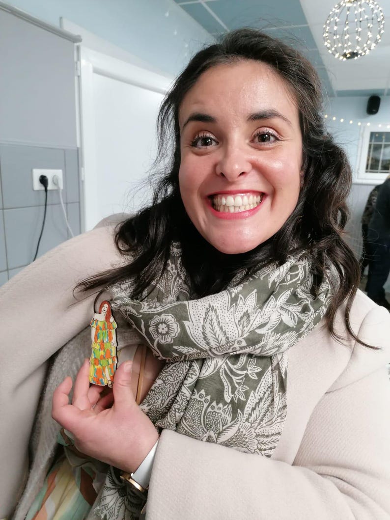 Pu&ograve; includere: Una donna indossa un cappotto bianco e una sciarpa verde con un motivo floreale e tiene in mano una piccola statuetta di legno di una donna con un vestito colorato. La statuetta &egrave; alta circa 10 cm.