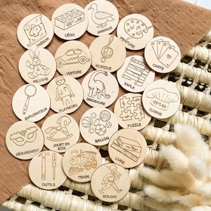May include: A set of 20 wooden circles with various images and French words on them. The images include a toy car, a dinosaur, a chef, a train, a soldier, a book, a ball, a puzzle, a rocking horse, a magnifying glass, a mask, a set of tools, a Lego brick, a stuffed animal, a musical note, a paint palette, a deck of cards, a stack of wooden blocks, and a duck. The words on the circles are "Tresor", "Lego", "Animaux", "Peluche", "Voiture", "Explorateur", "Deguisement", "Playmobil", "Peinture", "Carte", "Musique", "Kapla", "Dinosaure", "Jouet en Bois", "Ballon", "Puzzle", "Outils", "Train", "Livre", "Soldat", and "Cuisine".