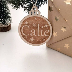 May include: A wooden Christmas bauble ornament with the name "Calie" engraved on it. The ornament is decorated with snowflakes and stars.