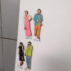 Pu&ograve; includere: Sagome di legno di quattro figure, due coppie, fissate su una superficie bianca. Una donna indossa un abito rosa e porta una borsetta, mentre l'uomo indossa una giacca blu e pantaloni arancioni. L'altra donna indossa un abito nero con dettagli floreali e l'uomo indossa una camicia verde e jeans. I nomi sono scritti sotto ogni figura.