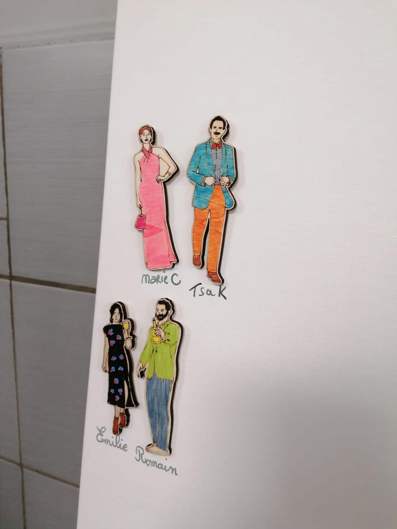 Pu&ograve; includere: Sagome di legno di quattro figure, due coppie, fissate su una superficie bianca. Una donna indossa un abito rosa e porta una borsetta, mentre l'uomo indossa una giacca blu e pantaloni arancioni. L'altra donna indossa un abito nero con dettagli floreali e l'uomo indossa una camicia verde e jeans. I nomi sono scritti sotto ogni figura.