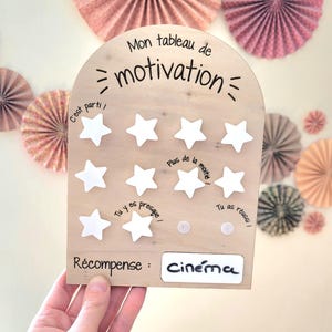 Könnte beinhalten: Eine Holz-Motivationskarte mit einer weißen Bogenform und dem Text "Mon tableau de motivation" oben. Die Karte hat acht weiße Sterne, die zum Verfolgen des Fortschritts verwendet werden können. Der Text "C'est parti!" steht neben dem ersten Stern, "Tu y es presque!" steht neben dem sechsten Stern, "Plus de la moitie" steht neben dem siebten Stern und "Tu as réussi!" steht neben dem achten Stern. Der Text "Récompense: Cinéma" steht unten auf der Karte.