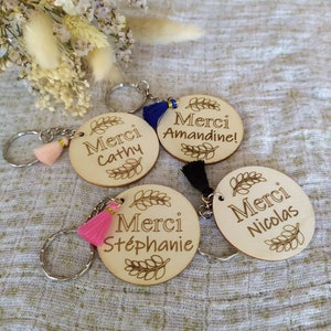 May include: Four wooden keychains with the word "Merci" and a name on each. The keychains are decorated with small tassels in pink and blue.