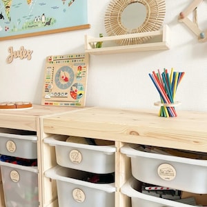 May include: A wooden shelf with a round mirror with a woven frame. Below the shelf are wooden storage bins with labels for toys, books, and other items. The bins are labeled with pictures of a train, a book, a robot, a picture, and a puzzle piece. The word "Jules" is written on the wall above the shelf.