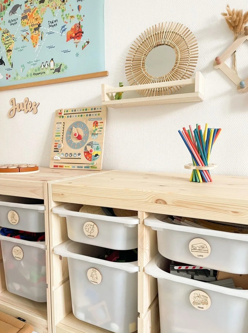 May include: A wooden shelf with a round mirror with a woven frame. Below the shelf are wooden storage bins with labels for toys, books, and other items. The bins are labeled with pictures of a train, a book, a robot, a picture, and a puzzle piece. The word "Jules" is written on the wall above the shelf.