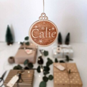 May include: A wooden Christmas bauble ornament with the name "Calie" inscribed on it. The ornament is decorated with white snowflakes and stars.