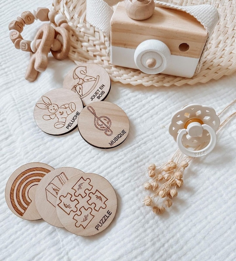 May include: Wooden baby toys with different themes, including a rocking horse, stuffed animal, music note, rainbow, house, and puzzle. The toys are on a white surface with a wooden camera and a pacifier.