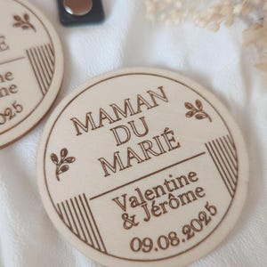 Può includere: Un cartello rotondo in legno con il testo "Maman du Marié" in un carattere decorativo. Sotto il testo si trova "Valentine & Jérôme" e la data "09.08.2025".