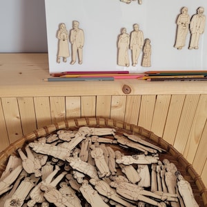 Pu&ograve; includere: Sagome di legno di persone in vari abiti, tra cui abiti da sposa, sono esposte su una lavagna bianca e in un cesto intrecciato. Matite colorate sono su una mensola di legno. Le sagome sembrano essere per un progetto fai-da-te.