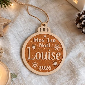 Può includere: Una decorazione natalizia rotonda in legno con un bordo in legno chiaro e un centro in legno scuro. Il testo riporta Mon 1er Noël, Louise e 2026, decorato con stelle e fiocchi di neve incisi. È presente un cordino in juta per appenderlo.