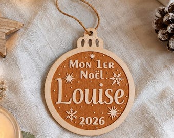 May include: A round wooden Christmas ornament featuring a light wood border and a darker wood centre. The text reads Mon 1er Noël, Louise, and 2026, accented with engraved star and snowflake designs. A jute twine loop is attached for hanging.