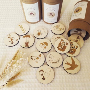 Peut inclure: Un jeu de mémoire en bois avec des illustrations variées, dont un oiseau, un écureuil, un cheval à bascule, une poule, un champignon, une famille, une paire de chaussures, une plante, une fille et un ours en peluche. Les tuiles sont dans une boîte en carton marron avec le texte "Jeu du Memory" sur le côté.