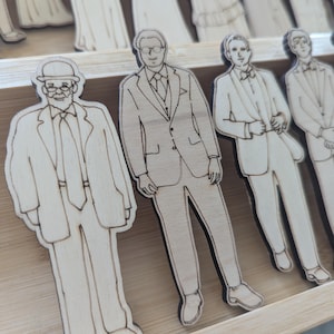 Pu&ograve; includere: Sagome di legno di uomini in abiti e smoking, probabilmente per un matrimonio. Le figure sono in legno chiaro con dettagli incisi, tra cui abiti, cravatte e papillon. Le sagome sono disposte in un vassoio di legno.