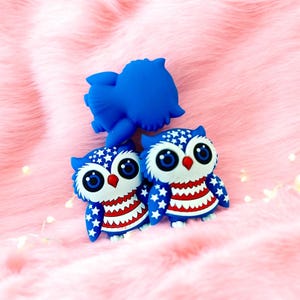 May include: Two blue owl figurines with white stars and red stripes. The owls have large eyes and are made of a soft, rubbery material.