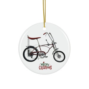 May include: Round white Christmas ornament featuring a vintage bicycle in burgundy and chrome. The bike has a high back seat and tall handlebars. The words "Merry Christmas" are written in red script at the bottom. A gold string is attached for hanging.