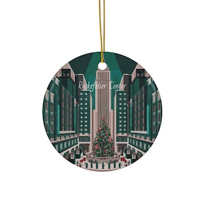 Può includere: Ornamento natalizio rotondo in ceramica con un'illustrazione stilizzata del Rockefeller Center a New York City. L'immagine mostra un albero di Natale decorato, edifici e il testo "Rockefeller Center". L'ornamento ha un cordino dorato per appenderlo.
