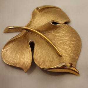 May include: A gold-toned leaf-shaped brooch with a textured surface. The brooch features three overlapping leaves and a curved stem, creating a layered design. The metal has a brushed finish, giving it a vintage look.