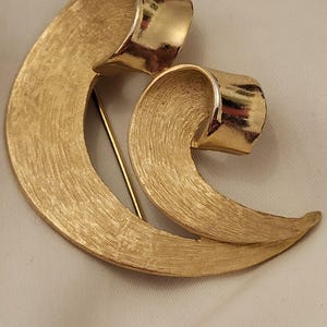 May include: A gold-toned brooch with a swirling, ribbon-like design. The brooch has a textured surface and a pin closure on the back. The design features two overlapping, curved elements, creating a sense of movement and elegance. A vintage jewelry piece.